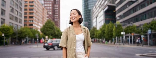 Venkovní záběr mladé usměvavé asijské ženy, stojící ve dne na ulici, rozhlížející se kolem, pózující šťastně. - Fotografie, Obrázek