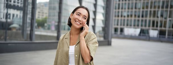 Uśmiechnięta, młoda Koreanka rozmawiająca przez telefon komórkowy i spacerująca po mieście. Szczęśliwa kobieta pozowanie na ulicy ze smartfonem. - Zdjęcie, obraz