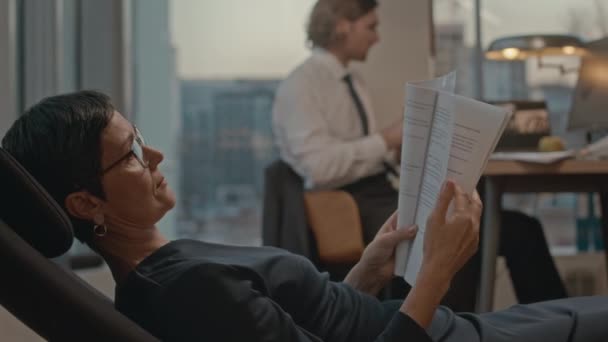 Ώριμη Καυκάσια επιχειρηματίας που γέρνει προς τα πίσω χαλαρώνοντας στην καρέκλα στο γραφείο της με πανοραμική παράθυρα διαβάζοντας το συμβόλαιο - Πλάνα, βίντεο