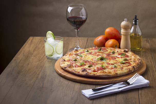 Lezzetli pizza hamuru, Mozzarella, domates, marjoram ve yeşil zeytinle yapılmış pizza. Tahtanın üzerinde. Napoli Pizzası. Bir bardak kırmızı şarap, zeytinyağı, biber öğütücü. 2 - Fotoğraf, Görsel