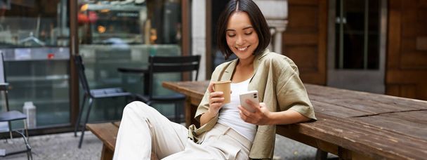 Stilvolles urbanes Mädchen sitzt mit ihrem Handy im Café, trinkt Kaffee und chattet und surft auf dem Smartphone in den sozialen Medien. - Foto, Bild