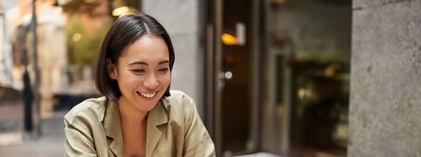 Uśmiechnięta młoda kobieta pracująca w kawiarni lub w miejscu pracy, siedząca z laptopem, ucząca się lub odrabiająca lekcje, wyglądająca na szczęśliwą. - Zdjęcie, obraz