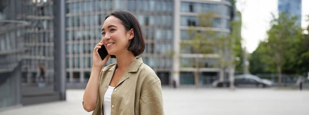 Вертикальний знімок молодої азіатської цифрової кочівниці, дівчина розмовляє на мобільному телефоні і гуляє на вулиці з ноутбуком. Молода жінка віддалений працівник йде в коворкінг
. - Фото, зображення