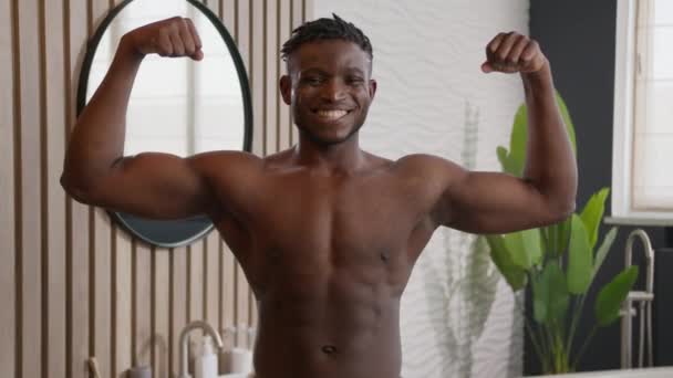 Izmos afro-amerikai férfi mosolygós sportos férfi testépítő kád törölköző a csípőn mutatja izmok bicepsz felemelése karok sport erős hatalom szexi etnikai srác meztelen törzs fürdőszobában ellátás a bőr test - Felvétel, videó