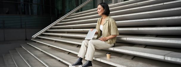 Jeune fille asiatique souriante avec ordinateur portable, se trouve sur la rue de la ville avec du café. Jeune femme fait des devoirs sur ordinateur tout en étant assis sur les escaliers de la rue, nomade numérique fonctionne à distance. - Photo, image