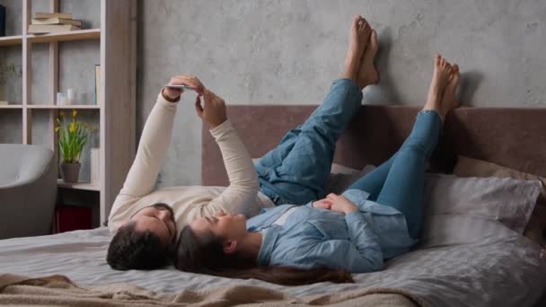 Kavkazský šťastný pár ležící vzhůru nohama společně na pohodlné posteli relaxovat s mobilním telefonem rolování sociální sítě nákup on-line nakupování uvolněný žena manželka a muž manžel volný čas v ložnici - Záběry, video