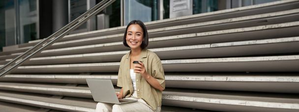 Vertical shot de sonriente chica estudiante, mujer asiática se sienta en las escaleras de la universidad campus y bebe café, hace su tarea en el ordenador portátil. - Foto, imagen