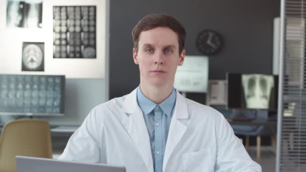Cintura hacia arriba POV de joven caucásico médico masculino asintiendo y gesticulando en la cámara sentado en el escritorio en la oficina médica moderna - Metraje, vídeo