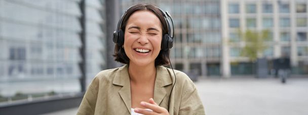 Zorgeloos Aziatisch meisje, lachend en glimlachend, koptelefoon dragend en lopend op straat. Outdoor shot van jonge vrouw luisteren muziek en op zoek gelukkig. - Foto, afbeelding