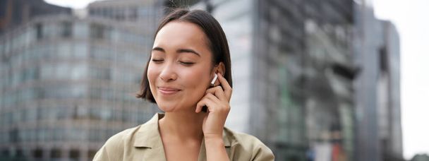 Vertikale Aufnahme einer lächelnden asiatischen Frau mit drahtlosen Kopfhörern, hört gerne Musik in Kopfhörern, hält ihr Handy in der Hand. - Foto, Bild