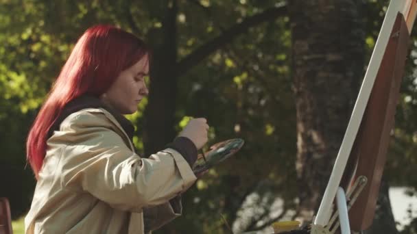 Zaostřené kavkazské dospívající dívka s jasnými rudými vlasy malování na plátně venku v parku osvětlené slunečním svitem - Záběry, video