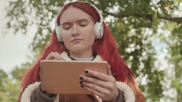 Мрійлива рожева дівчина-підліток в навушниках малює на графічному планшеті на відкритому повітрі - Кадри, відео