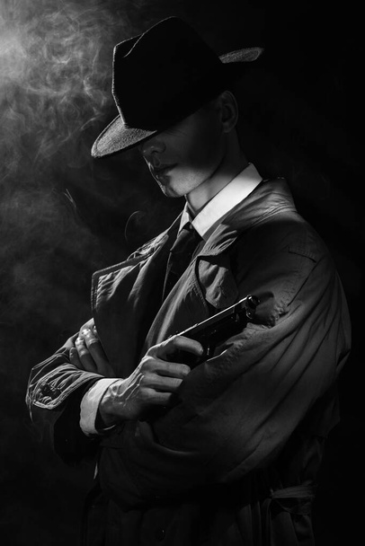 Темный силуэт человека в плаще и шляпе с пистолетом в руках в стиле криминального нуара. Драматический нуарный портрет в стиле детективов 1950-х годов - Фото, изображение