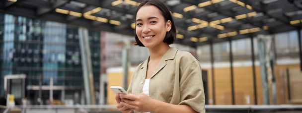 Technici. Mladá usměvavá asijská dívka drží smartphone, pomocí mobilní aplikace, zatímco stojí na ulici, navigace po městě. - Fotografie, Obrázek