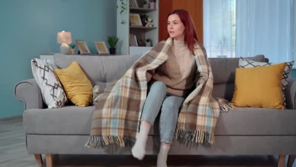 домохозяйка в теплом одеяле сидит на диване из-за холода дома, отсутствия отопления в доме - Кадры, видео