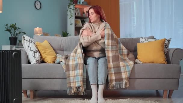 νεαρή γυναίκα είναι παγωμένη στο σπίτι λόγω έλλειψης θέρμανσης ή μη πληρωμής των χρεών για επιχειρήσεις κοινής ωφέλειας, μια νοικοκυρά ζεσταίνεται κάτω από ζεστή κουβέρτα κάθεται σε έναν καναπέ στο σπίτι - Πλάνα, βίντεο