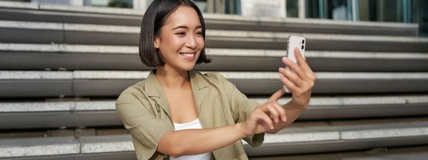 Retrato de asiático hermosa chica toma selfie en el teléfono inteligente. Mujer coreana joven se sienta en las escaleras al aire libre y hace fotos en el teléfono móvil. - Foto, Imagen