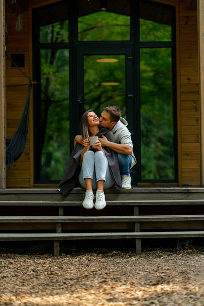 Une femme réfléchie s'assoit à la porte d'une maison en bois avec une tasse de thé avec son mari attentionné - Photo, image