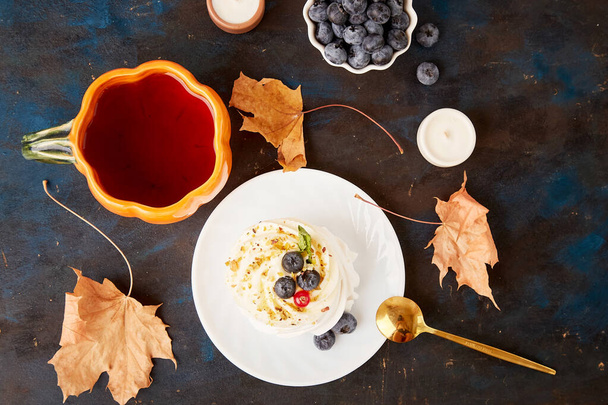 Herbst-inspirierte Pavlova mit Blaubeere und Kürbis-Teetasse zwischen trockenen Blättern auf rustikalem Hintergrund. - Foto, Bild