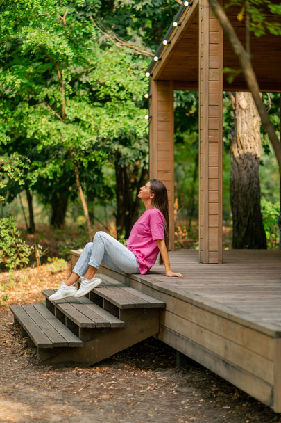 Вдумлива дівчина сидить з чашкою чаю на терасі свого дерев'яного котеджу в лісі вранці - Фото, зображення