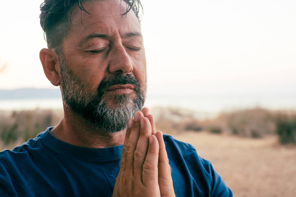 dospělý muž modlí a meditovat venku v relaxačním gestu s rukama sepjatýma a zavřenýma očima, portrét. Zen jako zdravý duševní vyrovnaný životní styl, mužští lidé. Blahobyt. Cvičení lidí - Fotografie, Obrázek