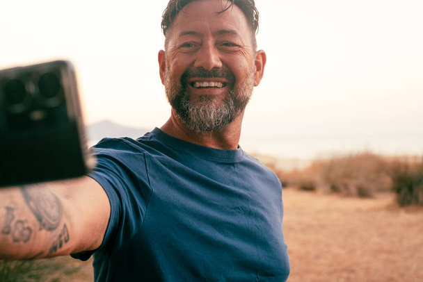Hombre feliz al aire libre tomando foto selfie usando teléfono inteligente móvil y sonriendo a la cámara. Compartir en línea la vida social. Personas y fotos de viajes. Cielo blanco fondo naturaleza escénica lugar campo - Foto, Imagen