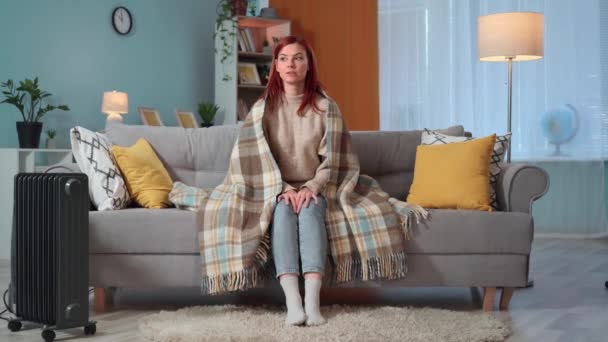 γοητευτική γυναίκα με ζεστή κουβέρτα ζεσταίνεται και μιλάει με την οικογένεια κάθεται στον καναπέ κοντά σε θερμάστρα στο σπίτι, κοιτάζοντας την κάμερα - Πλάνα, βίντεο