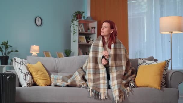 mladá žena zabalená v teplé přikrývce se snaží udržet teplo, protože zima uvnitř, zatímco sedí na gauči v nevytápěném bytě doma - Záběry, video