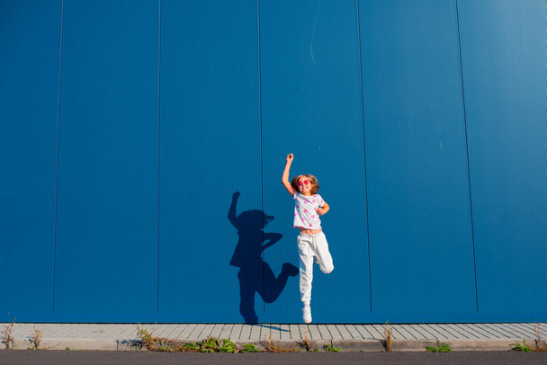 Joyful jong Europees blond meisje springen in een wit T-shirt en witte joggingbroek met roze hartvormige bril tegen een blauwe muur buiten - Foto, afbeelding