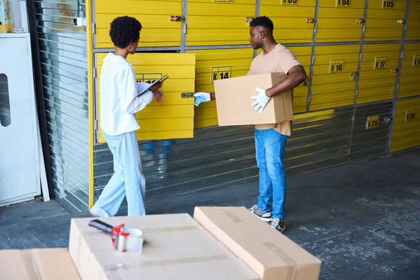 Afroameričan naloží kartónovou krabici do skladu, vedle manažerky s tabletem - Fotografie, Obrázek