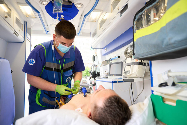 Arzt befestigt Elektroden an Saugnäpfen am Oberkörper eines Mannes im Krankenwagen, um ein Kardiogramm zu machen - Foto, Bild