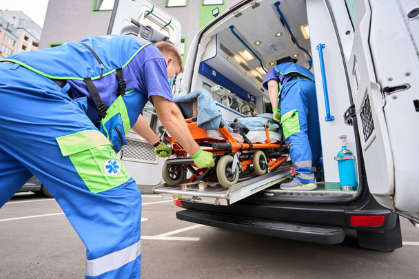 ambulancier dans un masque de protection décharge un patient d'une ambulance, son collègue est près du patient - Photo, image
