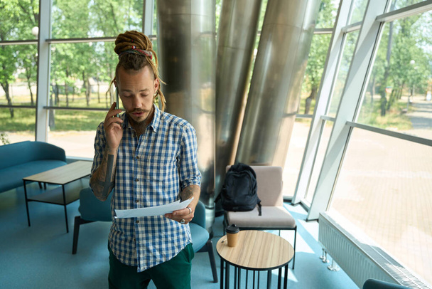 Homem com dreadlocks estuda documentos de trabalho e se comunica ao telefone, ele está em uma confortável área de coworking - Foto, Imagem