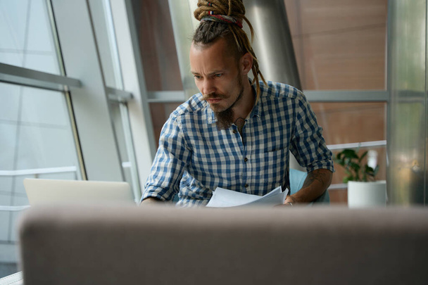 Hombre en una camisa a cuadros trabaja en un ordenador portátil en el área de coworking, que se encuentra en la vista - Foto, imagen