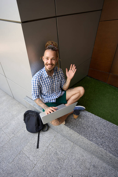 Informeller Typ mit Rucksack und Laptop sitzt auf einem Granitfelsen in der Nähe eines modernen Gebäudes - Foto, Bild