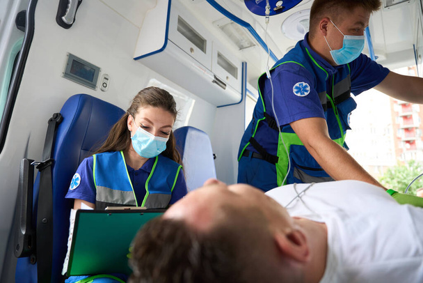Парамедики перевозят пациента в машине скорой помощи, медсестра заполняет анкету - Фото, изображение