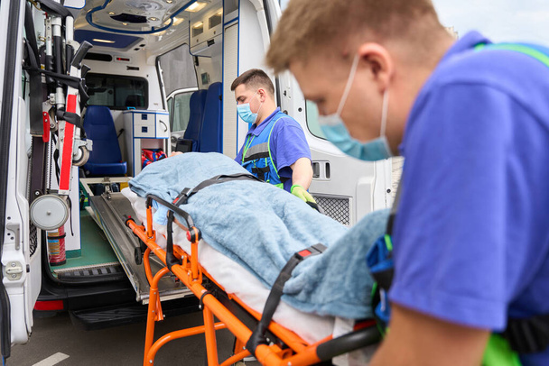 collègues masculins ambulanciers déchargent un brancard avec une victime d'une ambulance, dans la voiture il y a de l'équipement moderne - Photo, image