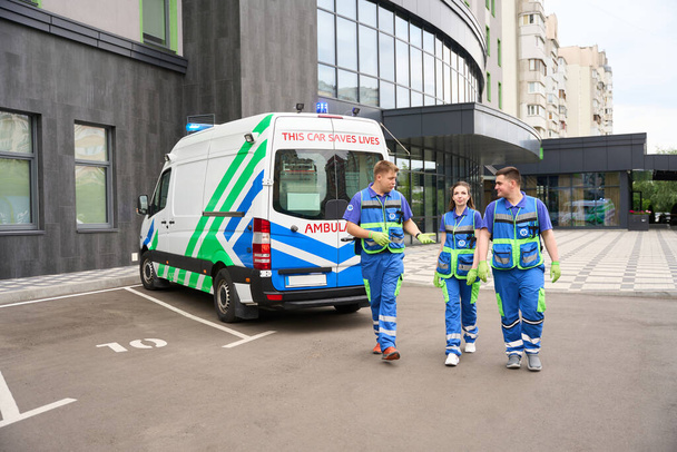 Tıp üniformalı insanlar modern bir binanın yakınındaki otoparkta yürüyorlar, yanında da bir ambulans var. - Fotoğraf, Görsel