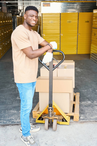 Joven hombre afroamericano en un almacén con un carro de carga, en el carro hay cajas de cartón con cosas - Foto, imagen