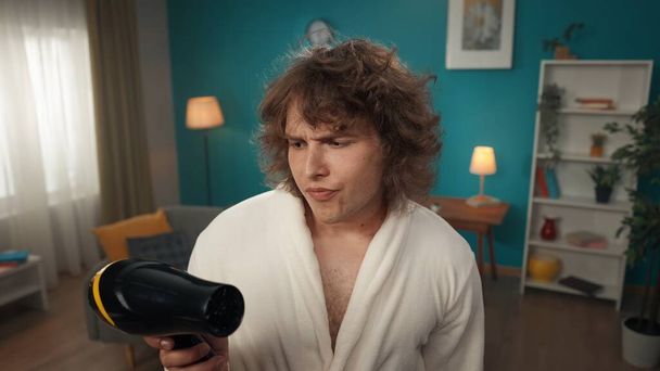A close-up of a young man with curly hair. He is dressed in a bathrobe and looks at the black hair dryer he holds in his hands. He portrays skepticism, distrust, doubt. He looks appraisingly. - Fotoğraf, Görsel