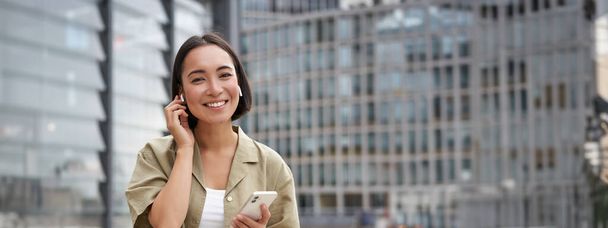Joven mujer asiática sonriente en auriculares inalámbricos, camina por la calle con teléfono inteligente y auriculares. - Foto, imagen