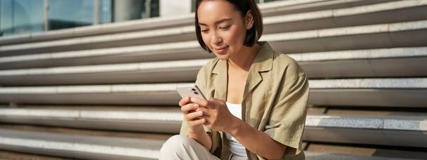 Люди и технологии. Улыбающаяся красивая азиатка, сидящая на лестнице в городе, держа в руках мобильный телефон. Девушка со смартфоном лежит снаружи. - Фото, изображение