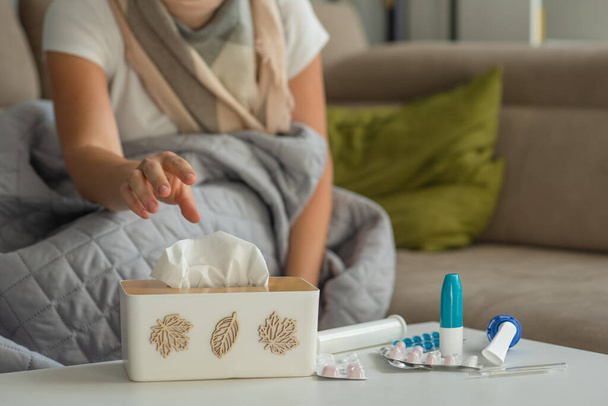 Leki na przeziębienia i papierowe serwetki są na stole, kobieta z objawami grypy i przeziębienia siedzi w tle i wyciąga serwetkę z pudełka. Skupienie się na lekach - Zdjęcie, obraz