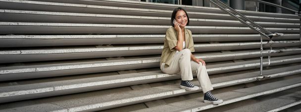 Smiling asian girl sits on stairs of building and talks on mobile phone, relaxing during telephone conversation. - Photo, Image