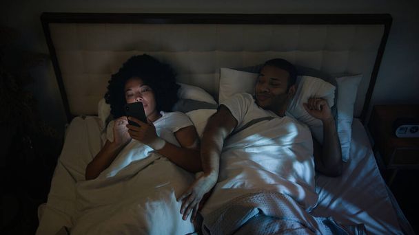 irytowany Afroamerykanin mężczyzna chłopak próbuje spać mąż zły push gadżet uzależniony żona internet uzależniona dziewczyna kobieta przewijanie telefon komórkowy social media w nocy w łóżku para problem rodzinny - Zdjęcie, obraz