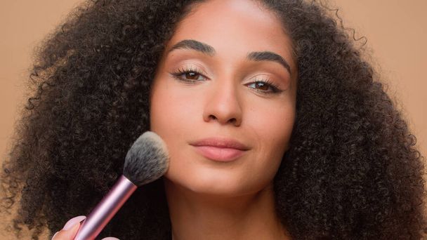 Close up portrait beige studio background beautiful African American woman with perfect smooth skin apply powder looking at camera attractive ethnic girl powdering face make-up cosmetics blush beauty - Photo, Image