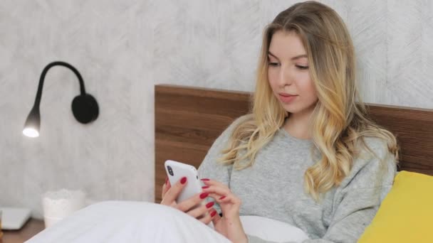 Szőke csinos nő ül az ágyon a szállodában vagy otthon, és beszélgetni a barátaival, vagy görgetés a szociális hálózatok okostelefonon. Emberek és technológiák koncepciója. Közelről. - Felvétel, videó
