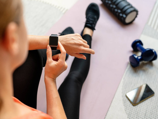 Vista ad alto angolo su sport smart watch con schermo bianco sul polso dell'atleta femminile si siede sul tappeto esercizio a casa. - Foto, immagini