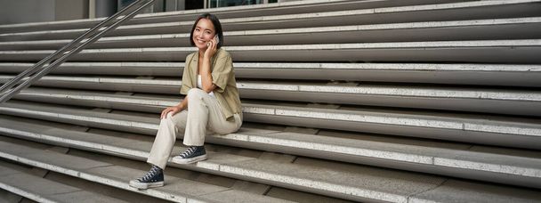 Усміхнена азіатська дівчина сидить на сходах будівлі і розмовляє на мобільному телефоні, розслабляючись під час телефонної розмови
. - Фото, зображення