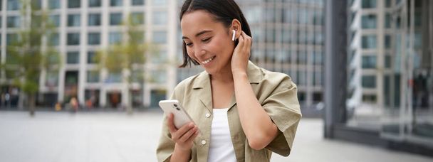 Sorrindo menina asiática escuta música em fones de ouvido sem fio, olha para o seu telefone, escolhendo música ou podcast. Jovem mulher chamando alguém, usando fones de ouvido. - Foto, Imagem